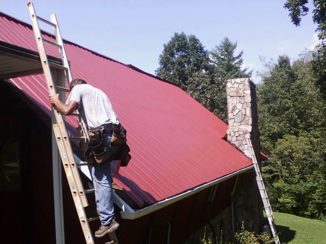Roofer on a ladder beside a red metal roof and stone chimney — Soffit & Fascia Repair by Roofing Brothers in West Oneonta